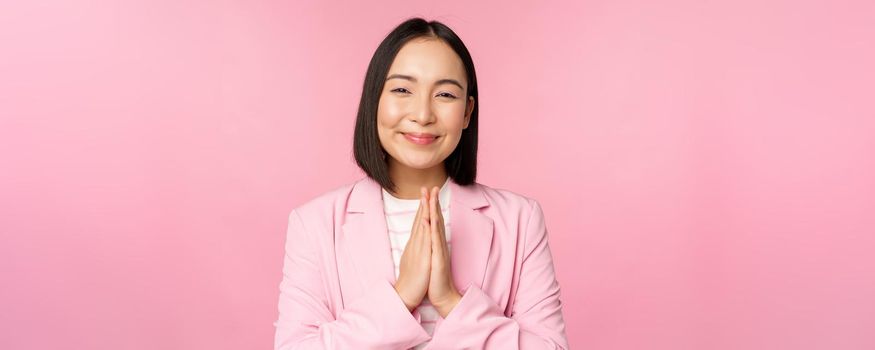 Thank you. Smiling asian saleswoman, corporate lady in suit thanking, holding hands in bag, gratitude gesture, standing over pink background.