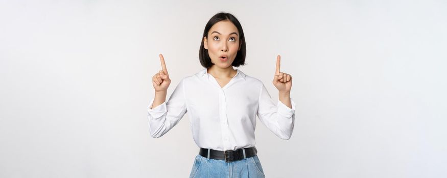 Enthusiastic asian business woman pointing, looking up with happy smiling face, showing company logo or banner, standing over white background.