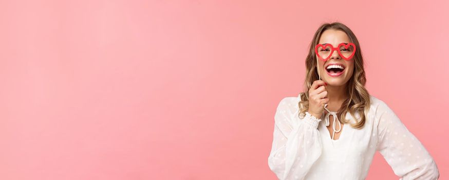 Spring, happiness and celebration concept. Close-up portrait of funny and carefree, beautiful caucasian woman with blond hair, white dress, holding heart-shaped glasses mask and laughing.