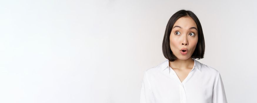 Close up portrait of young asian woman making surprised face, looking at upper left corner impressed, wow emotion, standing over white background.