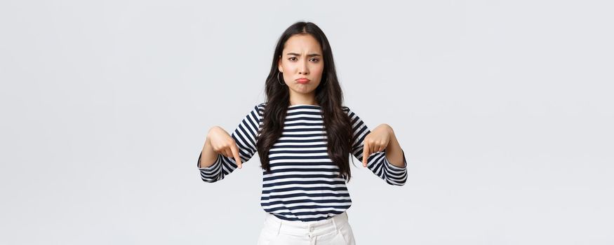 Lifestyle, beauty and fashion, people emotions concept. Disappointed upset young asian woman pouting and frowning displeased, pointing fingers down as complaining about product or service.