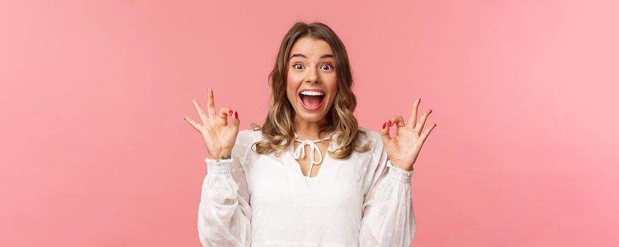 Portrait of excited and upbeat, cheerful blond girl in white dress dancing and showing okay signs with happy beaming smile, excited over music festival, rate awesome movie, recommend product.