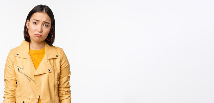 Portrait of sad korean woman sulking, frowning and looking upset, distressed frustrated face expression, standing gloomy against white background.