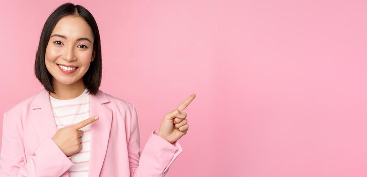Enthusiastic professional businesswoman, saleswoman pointing fingers right, showing advertisement or company logo aside, posing over pink background.