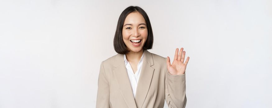 Friendly business woman, asian office lady waving hand and saying hello, hi gesture, standing over white background.