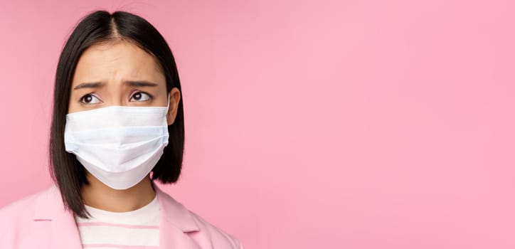 Close up portrait of sad, frowning asian businesswoman in medical face mask, looking left at copy space with disappointed, upset emotion, standing over pink background.