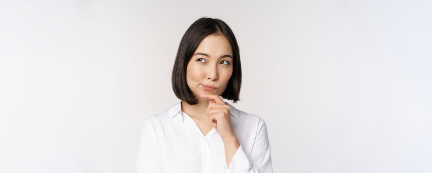 Close up portrait of asian woman thinking, looking aside and pondering, making decision, standing over white background.