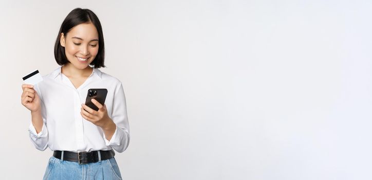 Online shopping concept. Korean woman holding credit card and looking at smartphone app, buying, order delivery in mobile phone application, standing over white background.