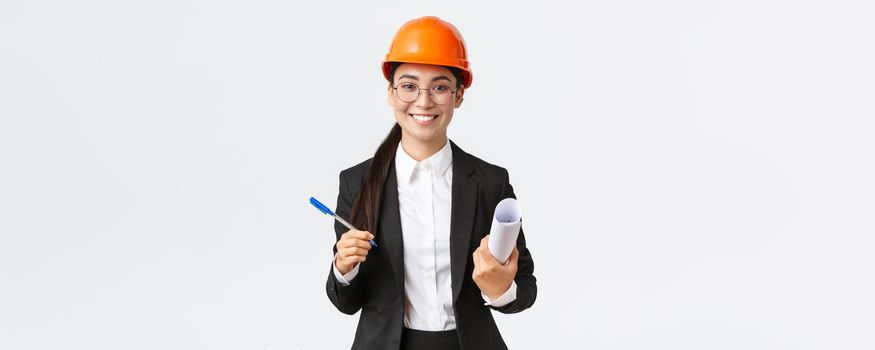 Smiling successful female entrepreneur, chief construction engineer in helmet and suit, holding blueprints and pen, signing contract with building supplier, white background.