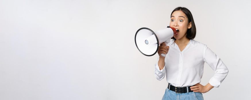 Image of young woman, korean activist, recruiter screaming in megaphone, searching, shouting at loudspeaker, standing over white background.