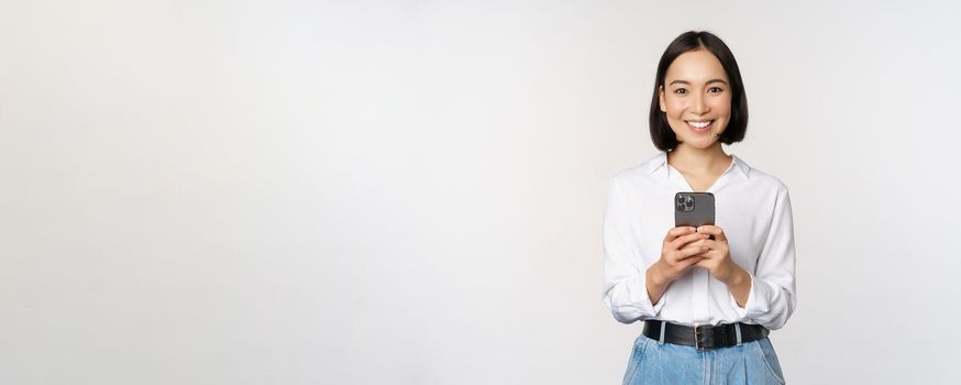 Stylish modern asian girl using mobile phone application, chatting on cellphone and smiling, standing in white blouse against studio background.