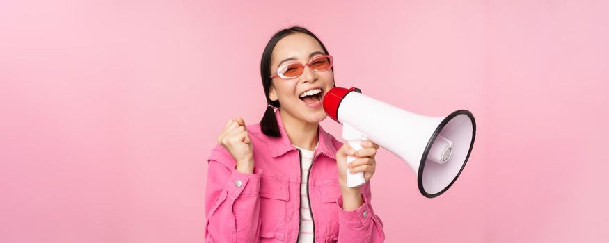 Attention, announcement concept. Enthusiastic asian girl shouting in megaphone, advertising with speaker, recruiting, standing over pink background.