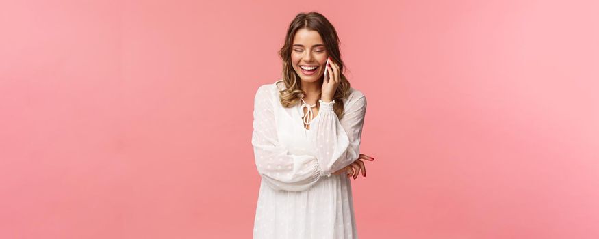 Communication and technology concept. Attractive caucasian female in white dress, blond short haircut, hold smartphone, talking on mobile phone and laughing, smiling carefree, pink background.