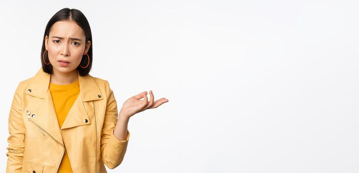 Image of frustrated korean woman, shrugging shoulders and raising hand, looking puzzled, cant understand smth, standing over white background.