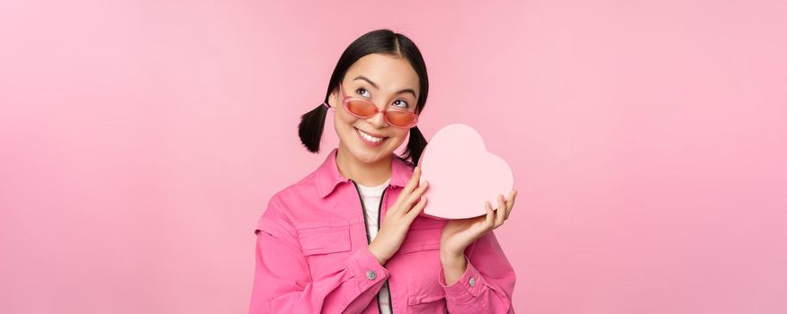 Image of stylish asian girlfriend in sunglasses, guessing whats inside gift box, heart-shaped present, standing over pink background.