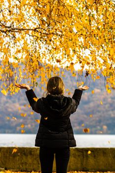 Back view of alone woman enjoying autumn, throwing fallen leaves on autumn alley. Autumn landscape, orange foliage in a park in Orsova, Romania, 2020