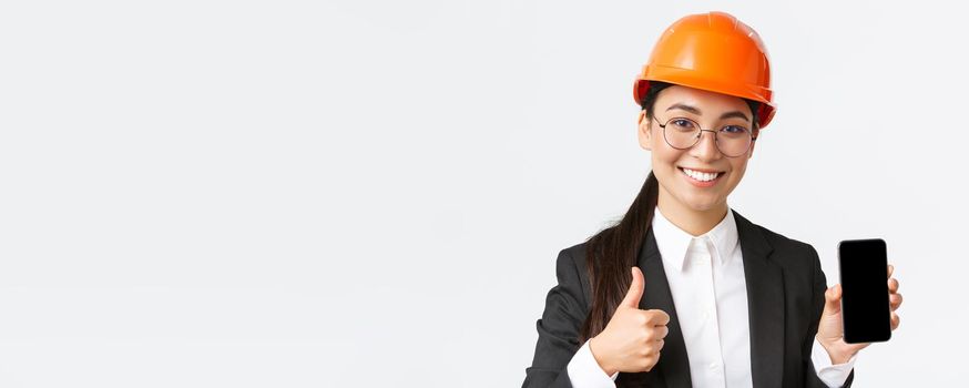 Close-up of professional smiling female engineer, construction manager in business suit and safety helmet, showing smartphone screen and thumbs-up in approval, white background.