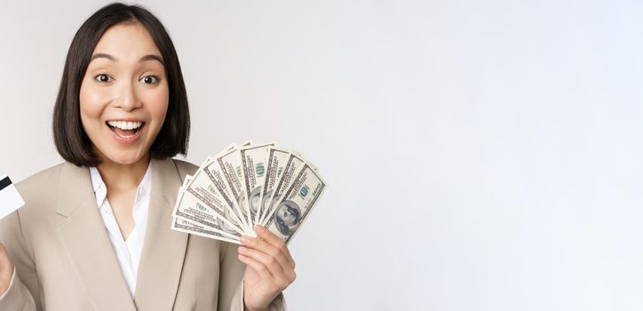 Enthusiastic young asian businesswoman, showing money dollars and credit card, standing over white background.