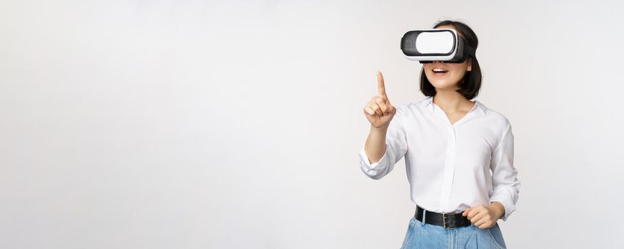 Amazed young asian woman using vr headset. Korean girl in virtual reality glasses pointing at smth and smiling, white background.