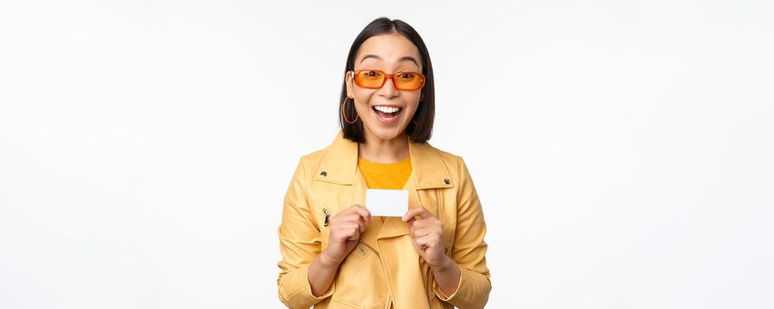 Portrait of beautiful modern asian girl in sunglasses, smiling happy, showing credit card, standing over white background. copy space