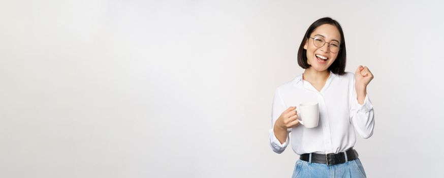 Happy dancing woman drinking coffee or tea from mug. Korean girl with cup, standing over white background.