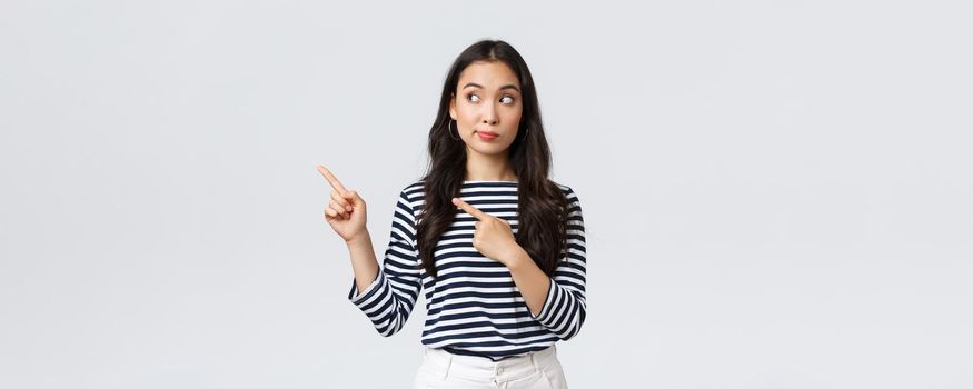 Lifestyle, beauty and fashion, people emotions concept. Curious and interested cute asian girl peeking and pointing fingers left at new promo banner with special discount in shop.