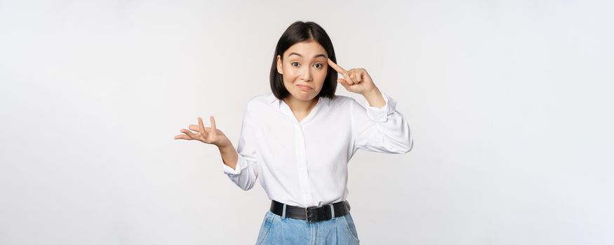 Are you insane. Portrait of frustrated young asian woman, roll finger near head and srugging, someone crazy or stupid, standing over white background.