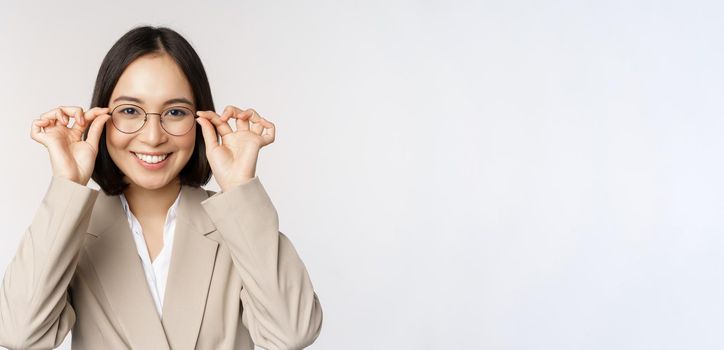 Smiling asian businesswoman trying new glasses, wearing eyewear, standing in suit over white background. Copy space