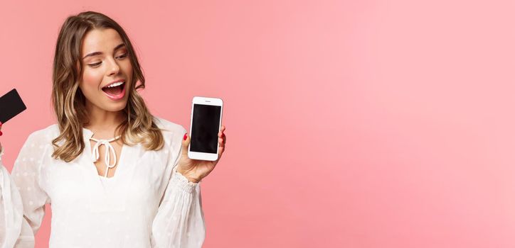 Finance, shopping and technology concept. Close-up portrait of excited good-looking blond girl in white dress, renew her spring wardrobe with buying clothes online via smartphone, hold credit card.