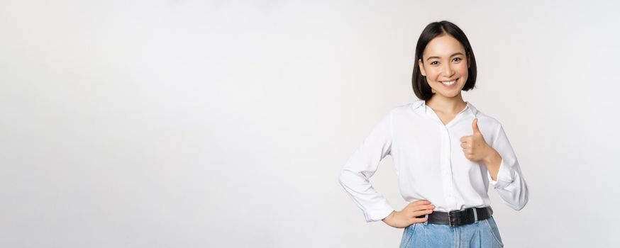 Image of confident asian woman showing thumb up in approval, recommending, like smth good, standing over white background.