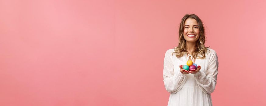 Holidays, spring and party concept. Portrait of tender, lovely blonde girl in white dress, holding Easter painted eggs, celebrating orthodox day, smiling cheerful share positivity, pink background.