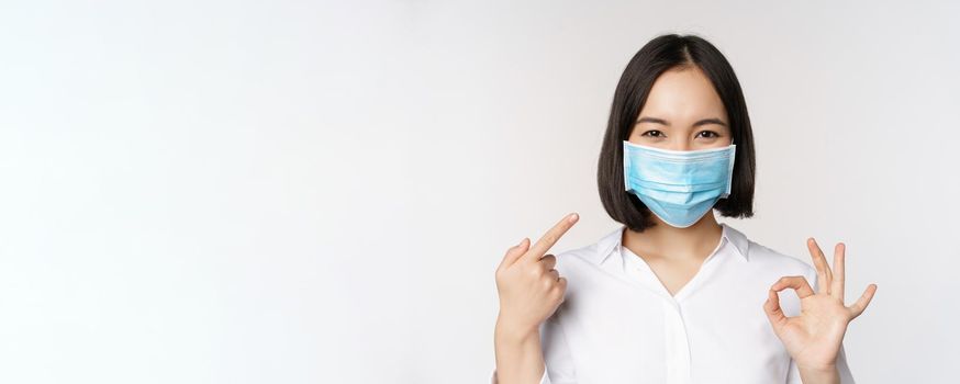 Portrait of asian girl in medical mask showing okay sign and pointing at her covid protection, standing over white background. Copy space