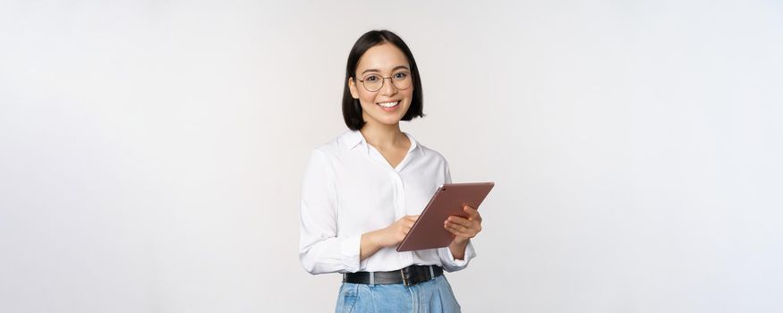 Image of young asian woman, company worker in glasses, smiling and holding digital tablet, standing over white background. Copy space