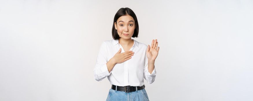 Image of cute young female office worker, asian girl student raising hand up and put palm on chest, name herself, introduce, making promise, standing over white background.