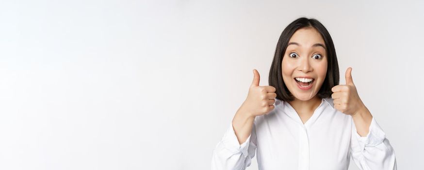 Enthusiastic asian girl showing thumbs up and smiling, pleased by something, recommending smth good, standing over white background.