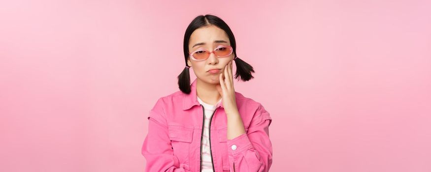 Image of sad asian girl sulking, touching her cheek, pouting disappointed, has toothache, standing over pink background.