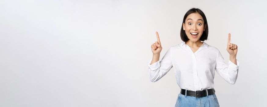 Enthusiastic business woman, asian female model pointing fingers up and smiling, making announcement, showing logo banner on top, white background.