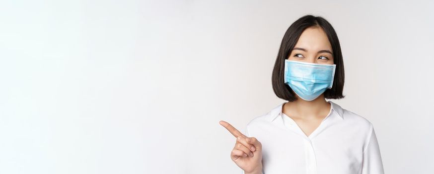 Portrait of cute asian woman in medical face mask, coronavirus protection, pointing finger left and looking intrigued at empty copy space, white background.