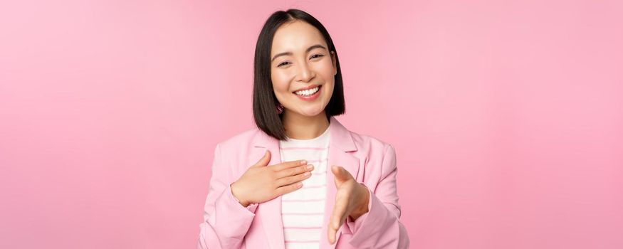 Portrait of smiling, pleasant businesswoman shaking hands with business partner, handshake, extending hand and saying hello, standing over pink background.