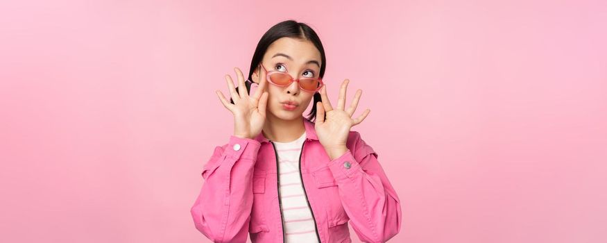 Close up of beautiful asian female model in stylish sunglasses, posing against pink background in trendy outfit, copy space.