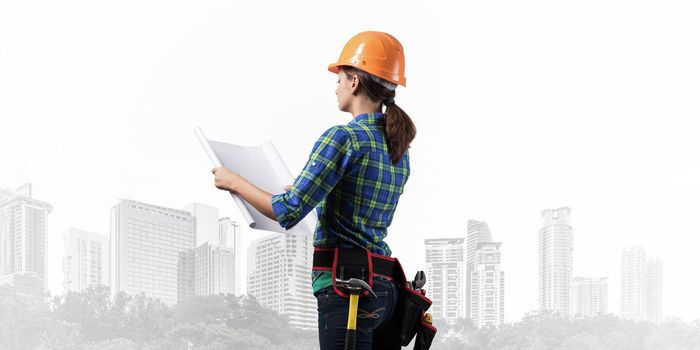 Female engineer in hardhat standing with technical blueprint. Side view of woman architect in checkered blue shirt inspecting construction on foggy metropolis background. Building project management