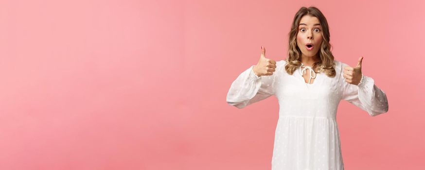 Portrait of amazed, excited blond young girl in white cute dress, open mouth say wow astonished and fascinated, show thumbs-up like and approve, beaing satisfied, pink background.