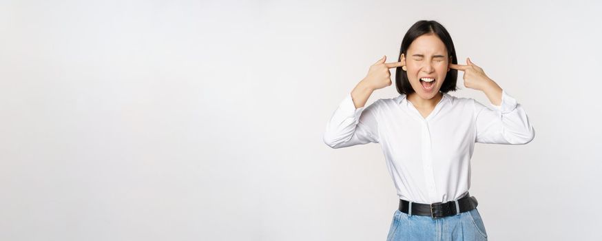 Annoyed asian woman feeling discomfort from loud annoying noise, close ears and eyes, standing over white background. Copy space