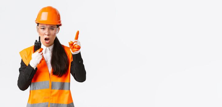 Serious-looking asian female industrial engineer, technician in safety helmet and uniform command construction team with walkie-talkie, explain or scolding someone with radio phone.