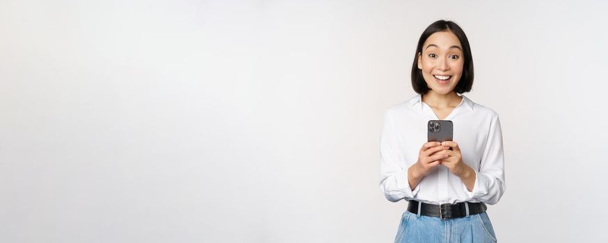 Excited asian woman smiling, reacting to info on mobile phone, holding smartphone and looking happy at camera, standing over white background.