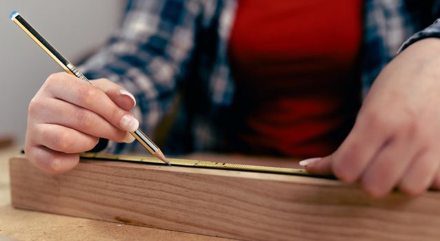 Detail of the hands of a young woman measuring a strip of wood for the manufacture of a piece of furniture in her small workshop, dressed in a blue checked shirt and red t-shirt. Young businesswoman handcrafting a piece of wood in her small business. Warm light indoors, background with wooden slats.