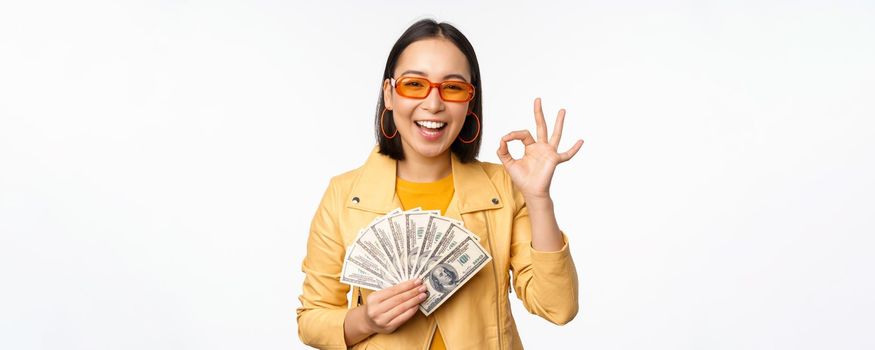 Microcredit and loans concept. Happy stylish korean girl showing okay sign, ok and money, dollars cash, standing in trendy clothes over white background.