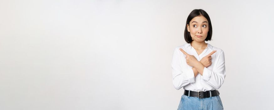 Indecisive asian woman pointing fingers sideways, pointing fingers and looking clueless, confused with choices, standing over white background.