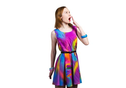 Surprised young female model with red hair looking up aside. Shocked woman hiding open mouth behind hand and feeling amazed. Elegant lady in colorful dress and bracelets against white background