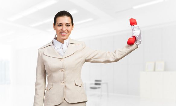 Woman keep at distance vintage red phone. Elegant operator in business suit posing with telephone in light office interior Hotline telemarketing and communication. Business assistance and support